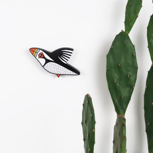 May include: A black and white wooden bird with orange and red accents, shaped like a puffin, is flying against a white background. A green cactus plant is in the background.