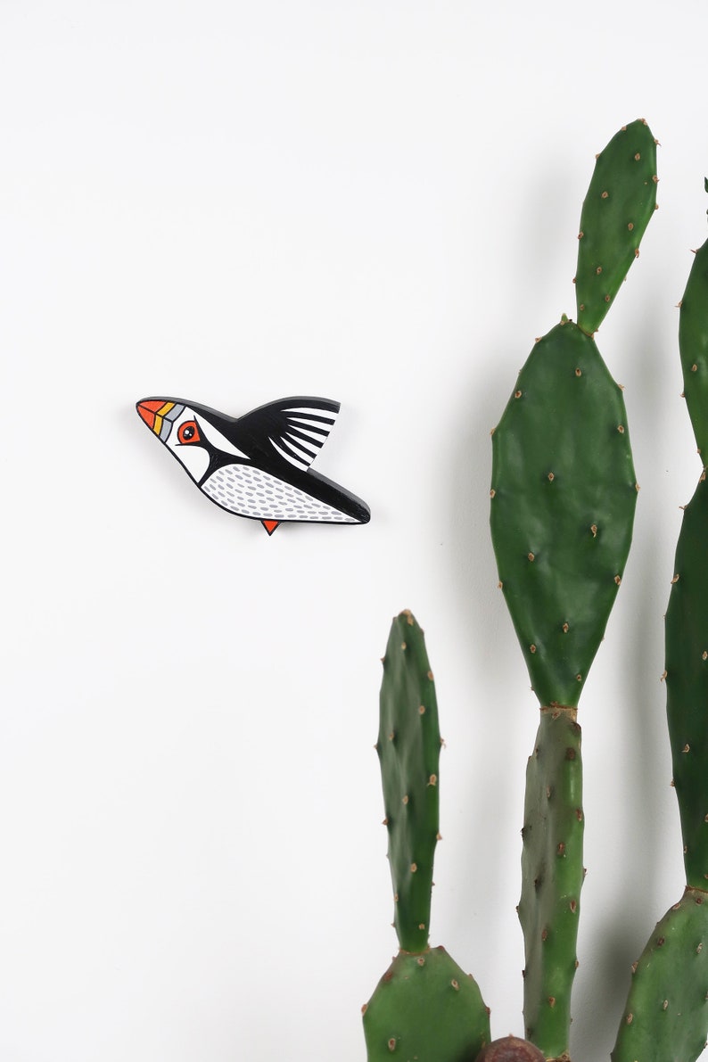 May include: A black and white wooden bird with orange and red accents, shaped like a puffin, is flying against a white background. A green cactus plant is in the background.