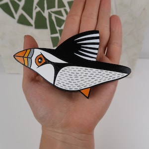 May include: A hand-painted wooden puffin ornament with black, white, orange, and gray details. The puffin is in flight with its wings spread out.