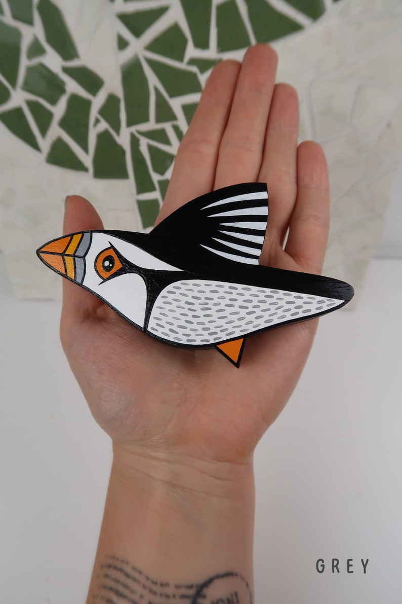 May include: A hand-painted wooden puffin ornament with black, white, orange, and gray details. The puffin is in flight with its wings spread out.