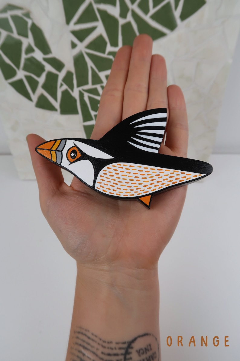 May include: A hand-painted wooden puffin ornament with black, white, orange, and gray details. The puffin is in flight with its wings spread out.
