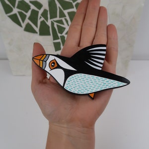 May include: A hand-painted wooden puffin ornament with black, white, orange, and turquoise accents. The puffin is in flight with its wings spread out.