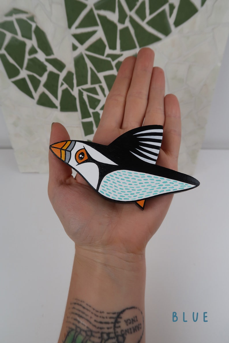May include: A hand-painted wooden puffin ornament with black, white, orange, and turquoise accents. The puffin is in flight with its wings spread out.