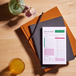 Op de afbeelding: Een witte dagelijkse planner met een groene kop die "Daily Planner" zegt. De planner ligt op twee notitieboekjes, een oranje en een zwarte. Er ligt een potlood op de notitieboekjes. Er staat een glas water met zaden en een vaas met paarse orchideeën op een houten oppervlak.