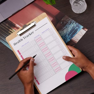 Op de afbeelding: Een witte gezondheidstracker op een houten clipboard, met een zwart potlood en een roze en groene cirkel. De tracker heeft secties voor wekelijkse maaltijdplannen, waterinname en work-out oefeningen.