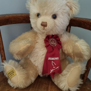 May include: A cream-coloured teddy bear named "Jeremy" with a red ribbon. The bear has dark eyes and a brown nose. It is sitting on a wooden chair. The bear has a tag on its foot.