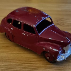 Pode incluir: Um carro de brinquedo bordô vintage com para-choque dianteiro prateado e pneus pretos. O carro clássico apresenta sinais de desgaste, sugerindo que é uma antiguidade. O carro de brinquedo está sobre uma superfície de madeira.