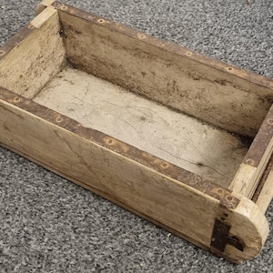 Könnte beinhalten: Eine rechteckige Holzkiste mit abgerundeten Enden und Metallbändern. Die Kiste hat ein verwittertes Aussehen mit sichtbarer Holzmaserung und gealterten Metallakzenten. Der Innenraum scheint unbemaltes Holz zu sein.