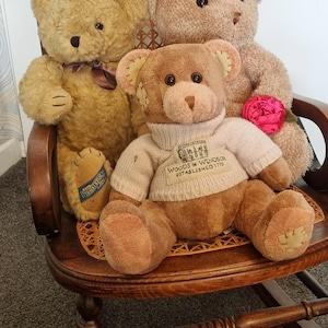 May include: Three teddy bears in shades of brown are arranged on a wooden rocking chair. The central bear wears a light-coloured sweater with the text "Woods of Windsor Established 1770". One bear holds a pink flower.