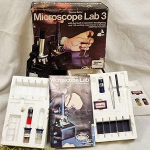 Pode incluir: Um conjunto vintage Thomas Salter Microscope Lab 3, incluindo um microscópio preto, um manual de instruções e vários acessórios de laboratório. A caixa apresenta uma ilustração do microscópio e o texto "Microscope Lab 3".