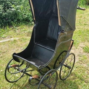 May include: A vintage-style black carriage with a high-sided body, a folding black fabric hood, and four thin metal spoked wheels. The interior features a black seat and backrest, with brown leather straps securing the frame and a handle at the rear.