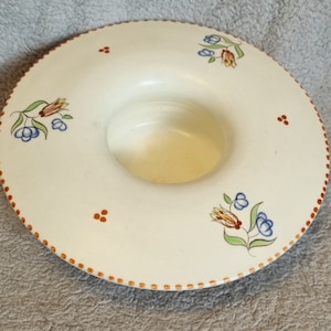 Puede incluir: Plato de cerámica de color crema con un borde ancho y poco profundo y un área central empotrada. El borde está decorado con pequeños puntos naranjas espaciados uniformemente y diseños florales en azul, naranja y verde. El plato tiene una estética vintage.