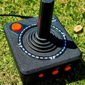 May include: A black Atari joystick controller with a black handle and orange buttons. The joystick has a textured surface and a white dotted ring around the base of the handle. The controller is on green grass.