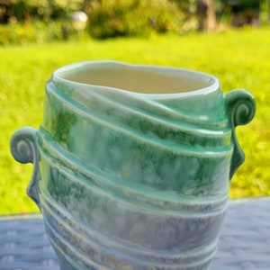 May include: A ceramic vase with a distinctive design. The vase is a mix of green, blue, and white hues. It has a wide opening and two decorative handles. The exterior has a textured, layered appearance, creating a visually appealing aesthetic.