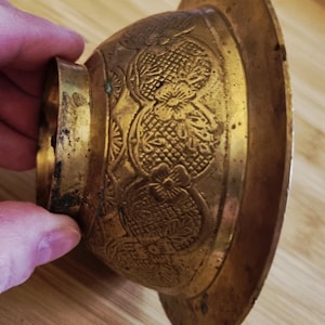 May include: A vintage brass bowl with intricate floral patterns and a wide, circular base. The bowl has a rounded body and a small, ring-shaped handle. The brass has a warm, aged patina, suggesting its antique nature.