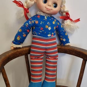 Könnte beinhalten: Eine Vintage-Stoffpuppe mit blonden Zöpfen, die mit roten Bändern gebunden sind. Die Puppe trägt ein blaues Oberteil mit Blumenmuster und weißer Spitzentrimmung sowie rot-blau gestreifte Hosen. Blaue Stoffschuhe vervollständigen das Outfit.