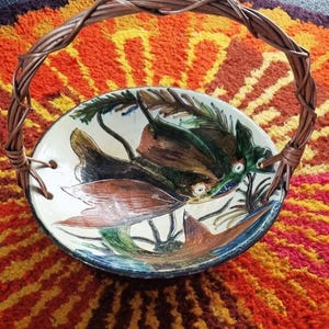 Könnte beinhalten: Dekorative Keramik-Servierschale mit geflochtenem Griff. Die Schale zeigt ein handgemaltes Design eines stilisierten Fisches in Grün-, Braun- und Orangetönen. Der Griff besteht aus verflochtenen braunen Zweigen. Die Schale steht auf einem farbenfrohen, strukturierten Teppich.