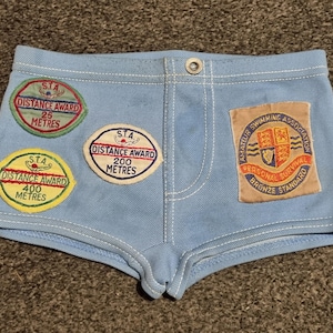 May include: Light blue swim trunks with multiple embroidered patches. Patches include "Distance Award" badges for 25, 200, and 400 meters, and an "Amateur Swimming Association" patch. The trunks have a button closure and white stitching.