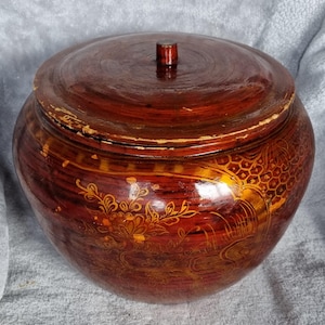 May include: A round, wooden container with a lid, painted in shades of red and brown. The container features intricate gold floral and geometric patterns. The lid has a small, cylindrical knob. The container is likely used for storage.