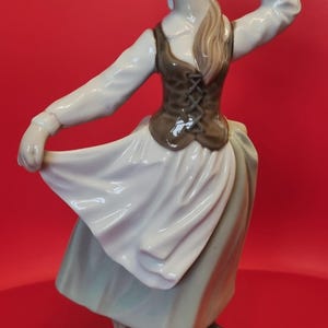 Puede incluir: Figura de porcelana de una joven con un vestido fluido, sosteniendo una flor. Lleva una blusa blanca, un chaleco marrón con cordones en la parte delantera y una falda larga. La figura está sobre una pequeña base texturizada, con fondo rojo.