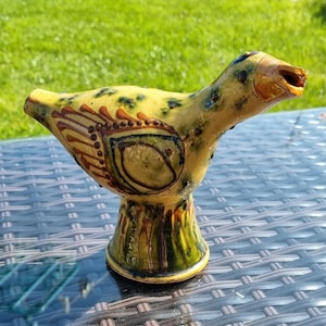 Puede incluir: Figura de pájaro de cerámica con cuerpo amarillo salpicado de puntos azul oscuro. El pájaro tiene patrones decorativos marrones y verdes en el lateral y una base verde acampanada. El pico está abierto y la figura está sobre una mesa de cristal.