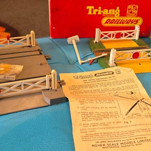 May include: A vintage Tri-ang Railways level crossing model set featuring a grey track section with white gates, a red storage box, a yellow road base, a small building, a Bird's Custard sign, and an instructional paper sheet with printed text.