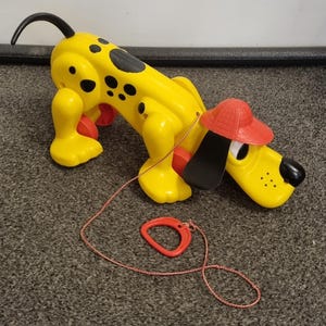 Può includere: Un giocattolo a forma di cane giallo da tirare, con macchie nere, una coda nera e un cappello rosso. Il cane ha un cordino rosso e un anello rosso. Il giocattolo è di plastica e ha ruote rosse.