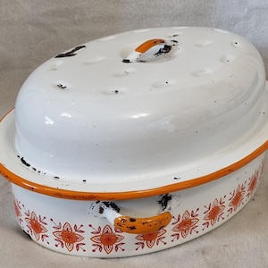 May include: Oval white enamel roasting pan with an orange rim and handles. The lid has small holes and an orange handle. The pan is decorated with orange floral patterns around the base. A vintage kitchenware item.