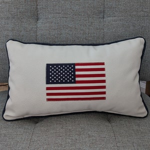 May include: A rectangular decorative pillow featuring the American flag design. The pillow has a white background with a navy blue border. The flag design includes red stripes and a blue square with white stars. The pillow is placed on a gray armchair.