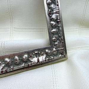 May include: A rectangular picture frame with a metallic silver finish and a raised, repeating relief pattern of acorns and oak leaves. The frame features a distressed, weathered texture with subtle dark red accents within the recessed areas.