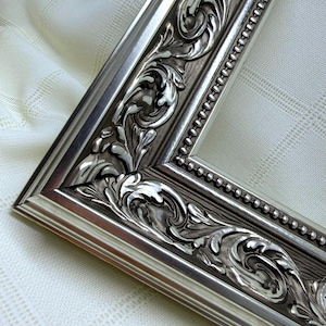 May include: A silver-toned picture frame featuring an ornate, raised scrollwork pattern along its border. The frame includes a beaded inner edge detail and a smooth, metallic finish that reflects light, creating depth across the intricate relief design.
