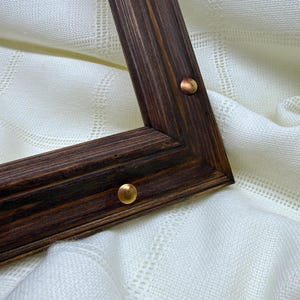 May include: A dark brown wooden frame with a visible wood grain texture and a stepped profile. Two small, round, metallic gold-toned decorative studs are embedded into the surface of the frame, positioned along the inner and outer edges.