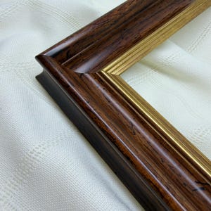 May include: A rectangular picture frame featuring a dark brown wood grain finish with a polished gold inner trim. The frame has a curved, moulded profile that creates a layered appearance, highlighting the contrast between the dark wood and gold.