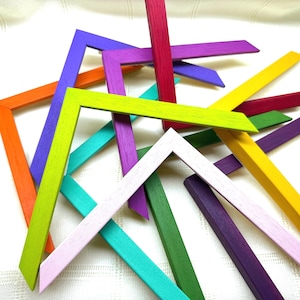 May include: A collection of L-shaped wooden picture frame corners in a variety of vibrant colours including lime green, orange, purple, magenta, yellow, teal, and light pink, all featuring a visible wood grain texture throughout the pieces.