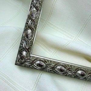 May include: A rectangular picture frame with a metallic silver finish and a weathered, antiqued texture. The frame features a repeating embossed pattern of oval shapes linked by curved, vine-like lines along its entire border.
