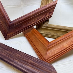 May include: Three wooden picture frame corners with distinct finishes. One corner features a deep reddish-brown wood grain, another displays a warm golden-brown tone, and the third shows a dark espresso brown hue. Each frame has a beveled edge.