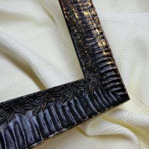 May include: A rectangular picture frame featuring a dark black finish with distressed gold highlights. The frame has a textured, ribbed inner edge and an outer border decorated with a repeating embossed leaf pattern along the entire length.