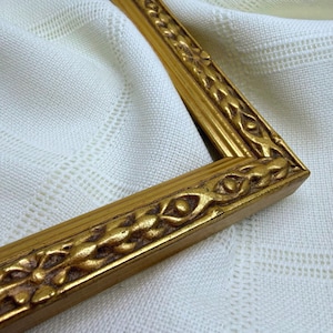 May include: A gold-coloured picture frame featuring a raised, repeating floral and vine-like relief pattern along its border. The frame has a metallic finish with darker recessed areas that highlight the intricate, sculpted details of the design.