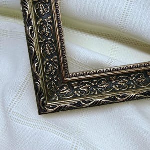 May include: An L-shaped corner section of a picture frame featuring a dark, recessed background with raised, metallic gold-toned relief patterns of scrolling leaves and vines, bordered by a thin, beaded edge detail along the inner perimeter.