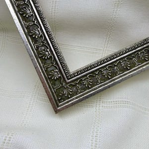 May include: A silver-toned picture frame corner featuring an intricate embossed floral pattern along the border. The frame has a metallic finish with dark recessed areas that highlight the raised leaf and vine details in a repeating design.