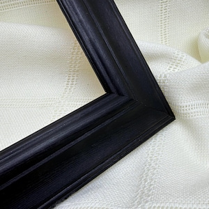 May include: A black picture frame with a mitered corner joint and a stepped profile. The frame features a visible wood grain texture throughout its surface, showcasing a dark finish that contrasts with the surrounding off-white fabric texture.