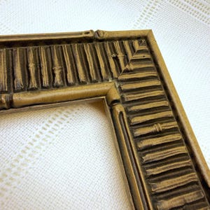 May include: A rectangular picture frame with a metallic gold finish and a dark brown antiqued wash. The frame features a raised, repeating bamboo-style texture along its border, with mitered corners and a deep, dimensional relief pattern.