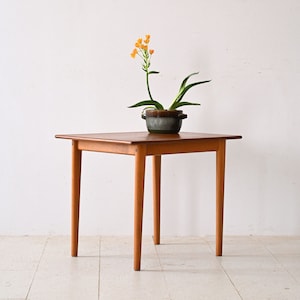 May include: A square wooden table with four legs. The table is made of light brown wood and has a smooth surface. A small potted plant with orange flowers is on the table.