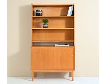 May include: A mid-century modern wooden bookcase with two doors and four shelves. The bookcase is made of light wood and has a simple design.