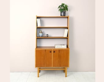 May include: A mid-century modern wooden bookcase with three shelves and two cabinet doors. The bookcase has a light wood finish and is standing on a white floor. A small potted plant is on the top shelf.