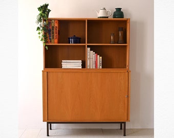 May include: A wooden cabinet with a hutch and sliding doors. The hutch has open shelves with books, a plant, and decorative items. The cabinet has a solid wood door and sits on black metal legs.