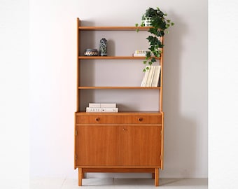 May include: A mid-century modern wooden bookcase with four shelves and a cabinet with two doors and a drawer. The bookcase has a plant on the top shelf and books on the lower shelves.
