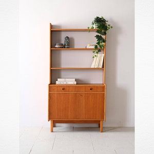 Puede incluir: Una estantería de madera de estilo moderno de mediados de siglo con cuatro estantes y un armario con dos puertas y un cajón. La estantería tiene una planta en el estante superior y libros en los estantes inferiores.