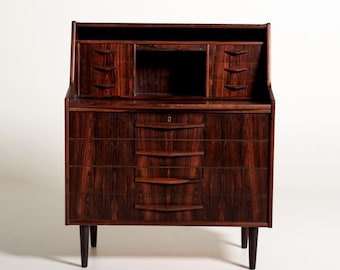 May include: A dark brown wooden bureau desk featuring a drop-down writing surface, four lower drawers with horizontal handles, and an upper section with two small drawers flanking a central open compartment, all supported by four tapered legs.