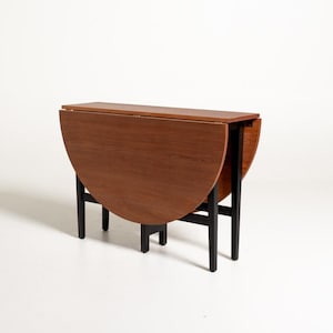 Puede incluir: Una mesa abatible con un tablero rectangular y dos alas laterales semicirculares que se pliegan hacia abajo. La mesa presenta un acabado de madera de color marrón cálido y se apoya en una estructura negra resistente con cuatro patas rectas.
